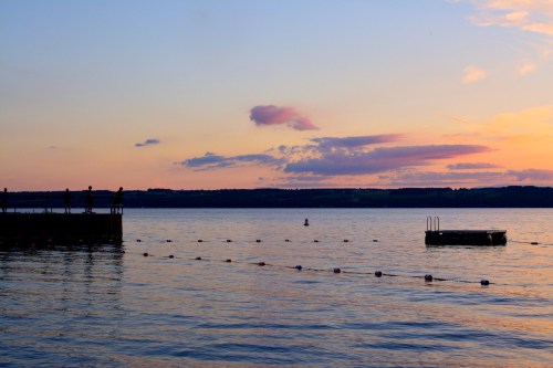 A serene sunset over a calm lake, with silhouettes of people on a pier and a floating platform in the water.