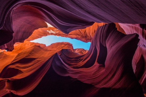 View looking up at the colorful, swirling rock formations of Antelope Canyon, with a bright blue sky above.