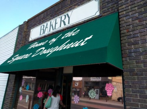 A bakery storefront with a green awning reading "Home of the Square Doughnut" and colorful flower decorations on the windows.
