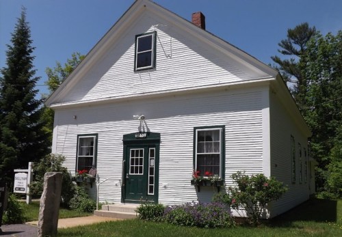 A white, two-story building with green accents, surrounded by greenery and flowers, under a clear blue sky.