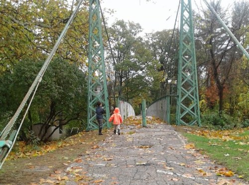 Two children walk on a leaf-covered path towards a green suspension bridge in a park during autumn.