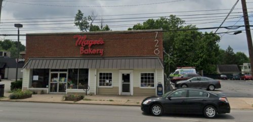 Exterior of Magee's Bakery, a brick building with a sign, surrounded by trees and parked cars on a cloudy day.