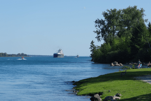 A large ship navigates a river, with small boats nearby and people sitting on the shore under clear blue skies.