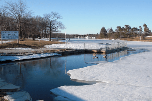 A peaceful park scene with a frozen lake, a dock, and snow-covered ground under a clear blue sky.