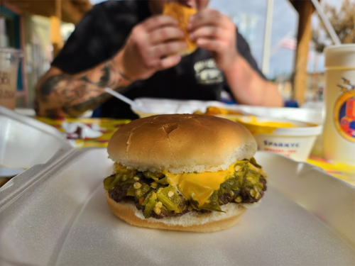 A close-up of a cheeseburger with pickles on a takeout container, with a person eating in the background.