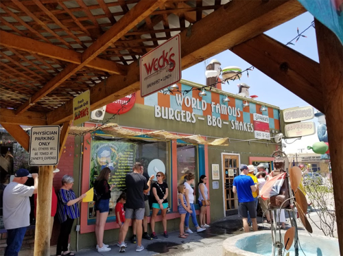 A busy restaurant exterior with a sign reading "World Famous Burgers - BBQ - Shakes" and a line of customers waiting outside.