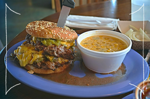 A large cheeseburger with multiple patties and melted cheese, served with a side of creamy corn soup on a blue plate.