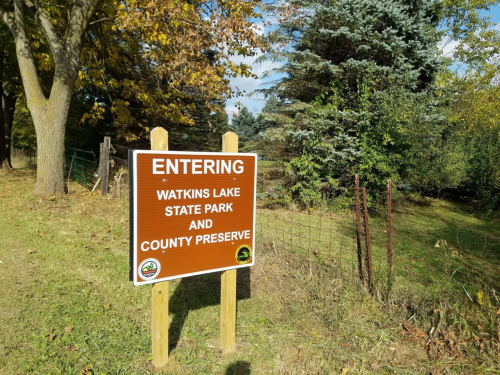 Sign marking the entrance to Watkins Lake State Park and County Preserve, surrounded by trees and greenery.