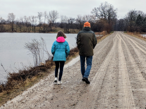 A man and a girl walk along a gravel path by a lake on a cloudy day, surrounded by bare trees.