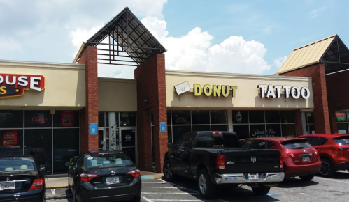 A storefront featuring a "Donut" shop and a "Tattoo" parlor, with parked cars in front and a blue sky above.