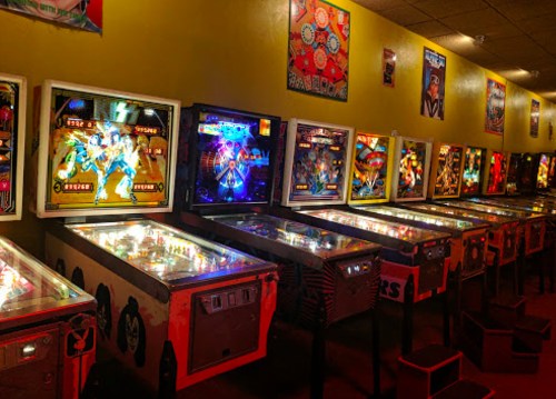 A row of colorful pinball machines in a retro arcade, illuminated under warm lighting.