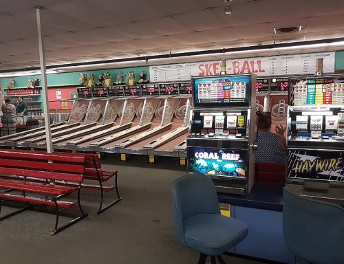 A retro arcade featuring several skee-ball lanes, a slot machine, and empty seating in a colorful setting.