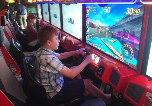 A boy plays a racing arcade game, focused on the screen, with colorful graphics and other players in the background.