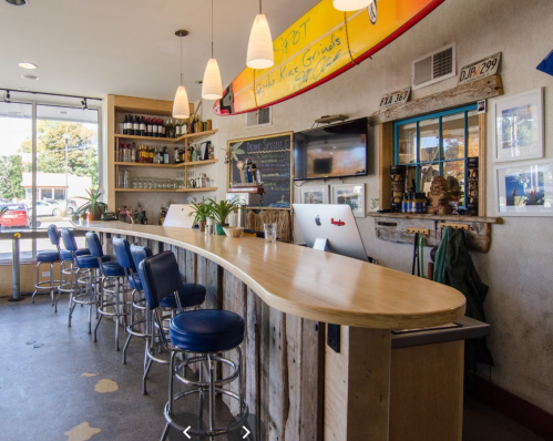A cozy bar with a curved wooden counter, blue stools, and surfboards on the wall, creating a relaxed atmosphere.