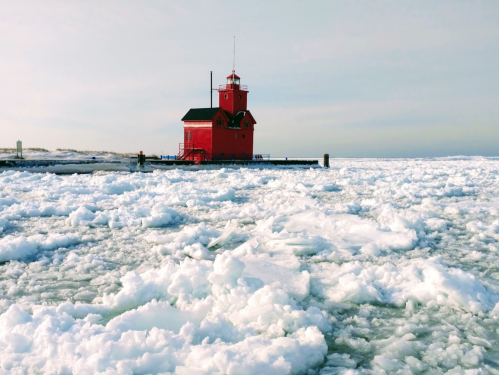 A red lighthouse stands on a pier surrounded by ice and snow, with a calm sky in the background.