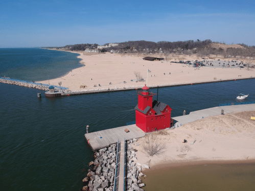 A red lighthouse on a pier by a sandy beach and calm water, with trees and buildings in the background.