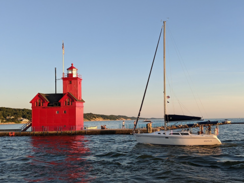 A red lighthouse stands by the water as a sailboat approaches during sunset.