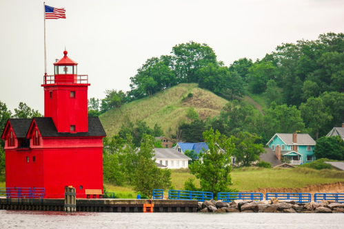 A red lighthouse stands by the water, surrounded by green hills and colorful houses under a cloudy sky.