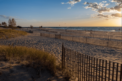 A serene beach at sunset, with sand dunes, wooden fences, and gentle waves under a colorful sky.