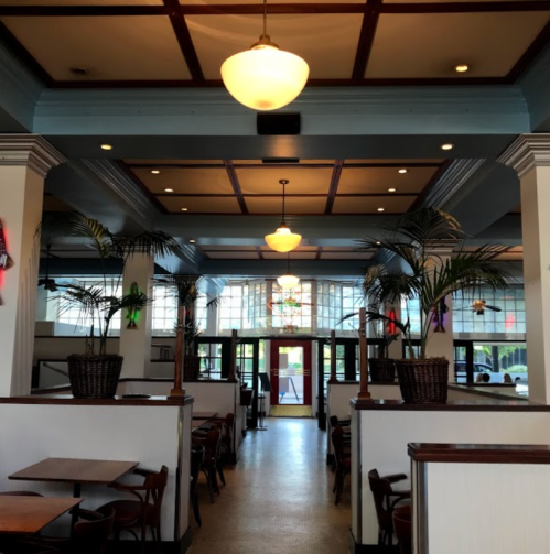 Interior of a restaurant featuring wooden tables, plants, and decorative lighting, with a bright entrance in the background.