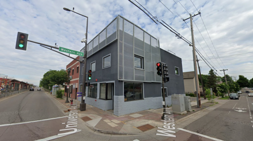 A modern building at the corner of Western Ave and University St, with traffic lights and a cloudy sky above.