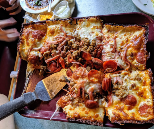 A tray of pizza topped with pepperoni, sausage, and melted cheese, served with a spatula on a table.