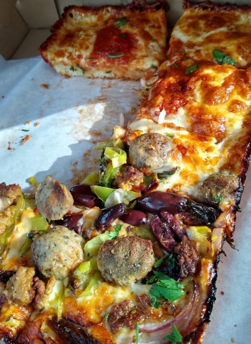 A close-up of a rectangular pizza topped with meatballs, olives, peppers, and melted cheese, with a slice removed.