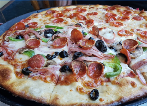 A close-up of a pizza topped with pepperoni, ham, green peppers, black olives, and melted cheese on a golden crust.
