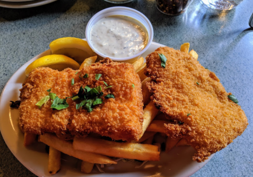 Two breaded fish fillets on a bed of fries, garnished with parsley, served with a side of tartar sauce and lemon wedges.