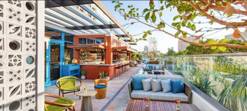 A vibrant outdoor lounge area with colorful furniture, greenery, and a bar, set in a sunny, modern environment.