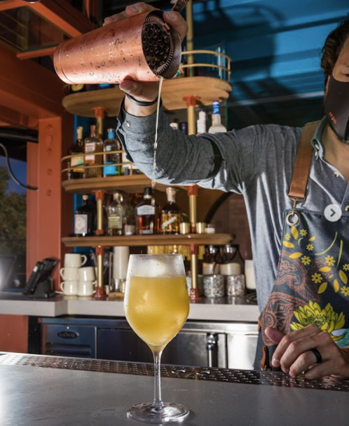 A bartender pours a drink from a shaker into a glass, with shelves of bottles in the background.