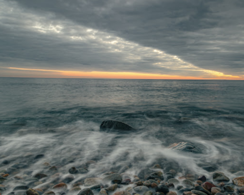 A rocky shoreline at sunset, with waves gently lapping against the stones under a cloudy sky.