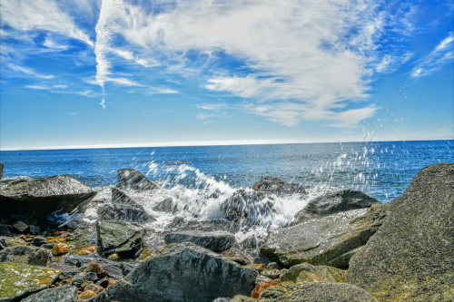 Waves crashing against rocky shore under a bright blue sky with scattered clouds.