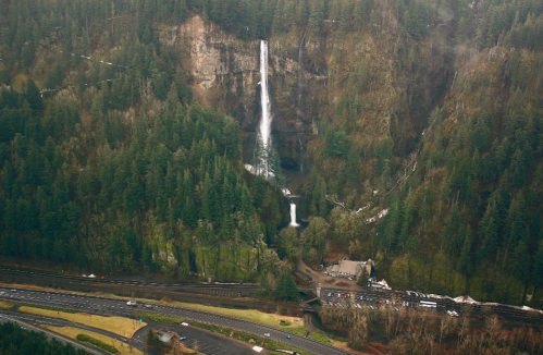 Aerial view of a lush green landscape featuring two waterfalls surrounded by dense forest and a winding road below.