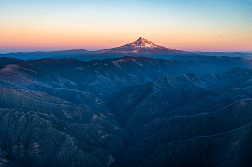 A panoramic view of a snow-capped mountain at sunset, surrounded by rolling hills and valleys.