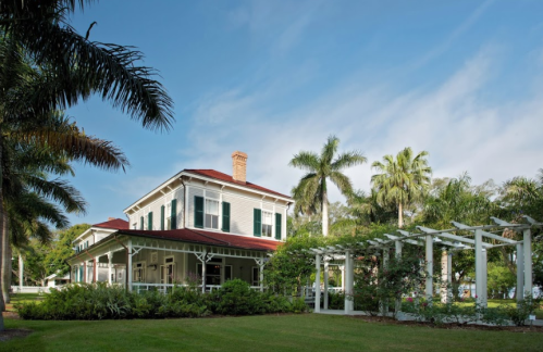 A charming two-story house with a red roof, surrounded by lush greenery and palm trees under a blue sky.