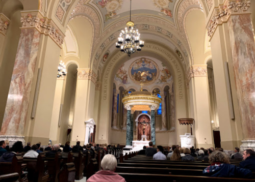 Interior of a church with a congregation seated, ornate architecture, and a central altar featuring a statue.