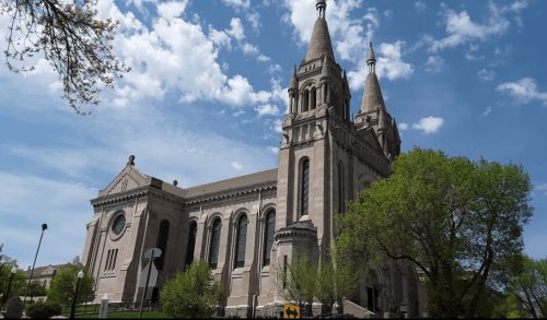 Historic church with twin spires, surrounded by greenery and under a blue sky with scattered clouds.