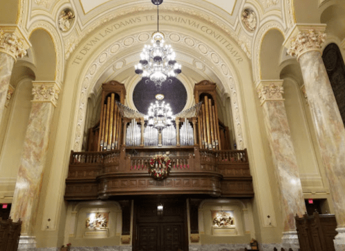 Interior of a grand hall featuring a large pipe organ, ornate chandeliers, and decorative architectural details.