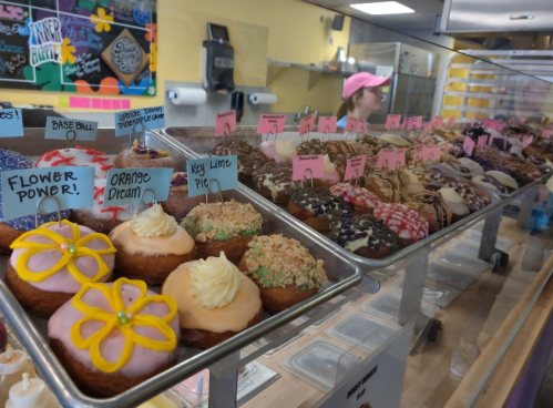 A display of colorful donuts with various toppings and flavors, with a staff member in the background.
