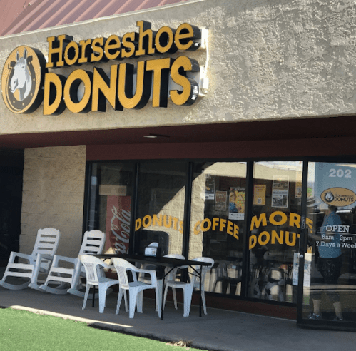 Exterior of Horseshoe Donuts shop with a sign, outdoor seating, and large windows displaying menu items.