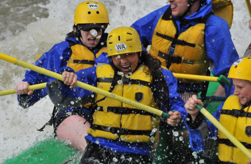 A group of people in yellow helmets and life jackets joyfully rafting through splashing water.