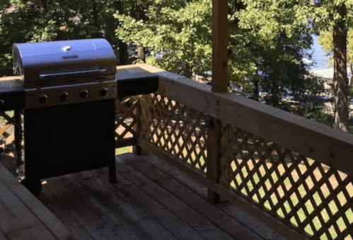 A stainless steel grill on a wooden deck surrounded by trees and a view of a lake in the background.