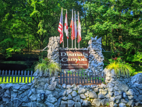 Sign for Dismals Canyon, featuring flags and surrounded by a stone wall and greenery.