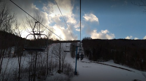 A ski lift ascends a snowy slope, surrounded by trees and a cloudy sky.