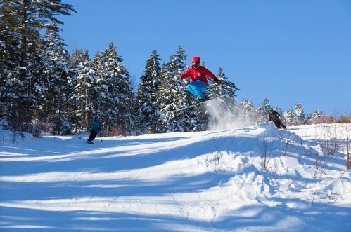 A skier jumps off a snow-covered slope, with two others skiing nearby in a winter landscape.