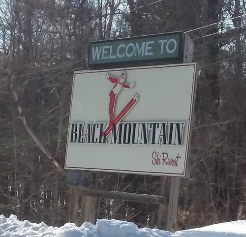 Welcome sign for Black Mountain Ski Resort, featuring a skier graphic and surrounded by snow-covered trees.