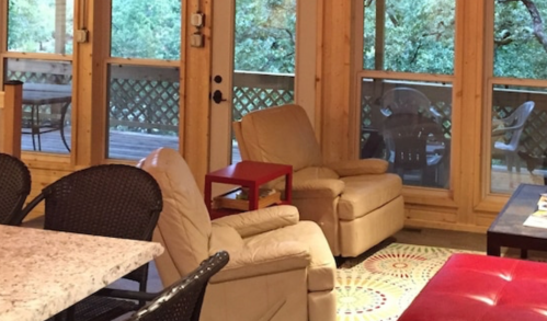 Cozy living space with two beige chairs, a red table, and large windows overlooking a deck and greenery.