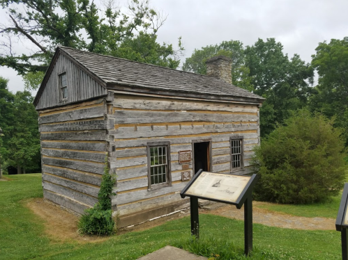 A rustic wooden cabin with a stone chimney, surrounded by greenery and a historical information sign in front.