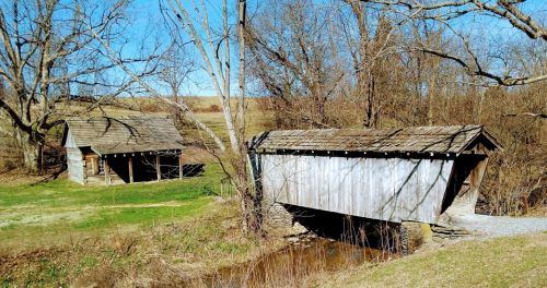 A rustic covered bridge next to a small wooden cabin, surrounded by bare trees and a clear blue sky.